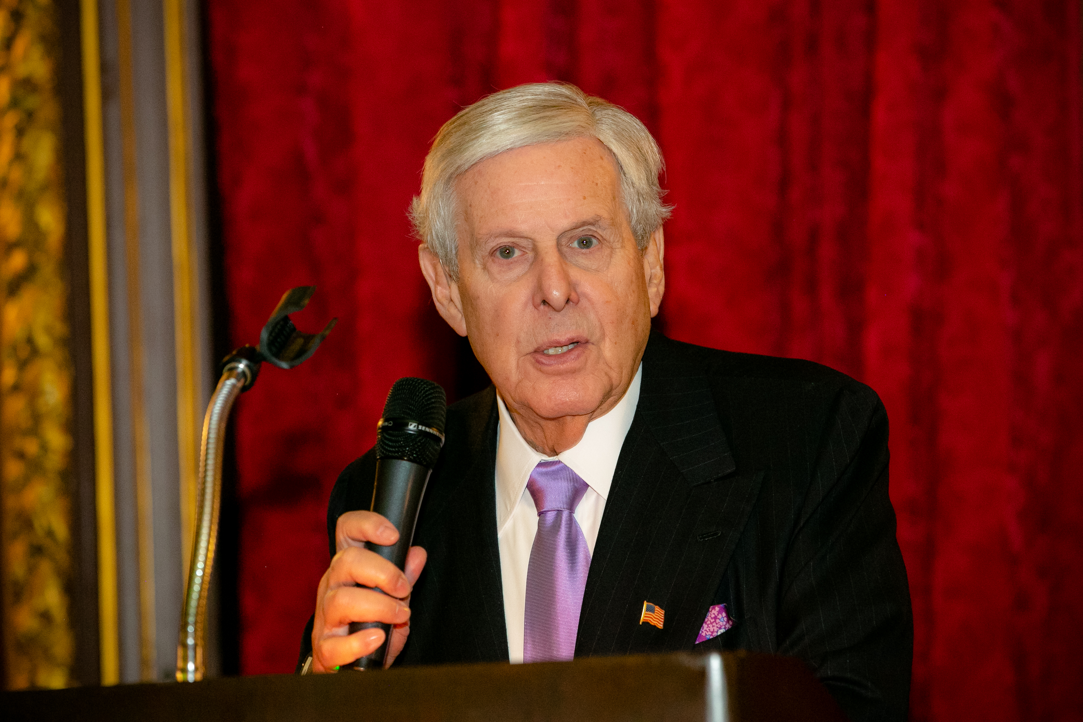 A man making a speech wearing a black suit with a purple tie and holding a microphone